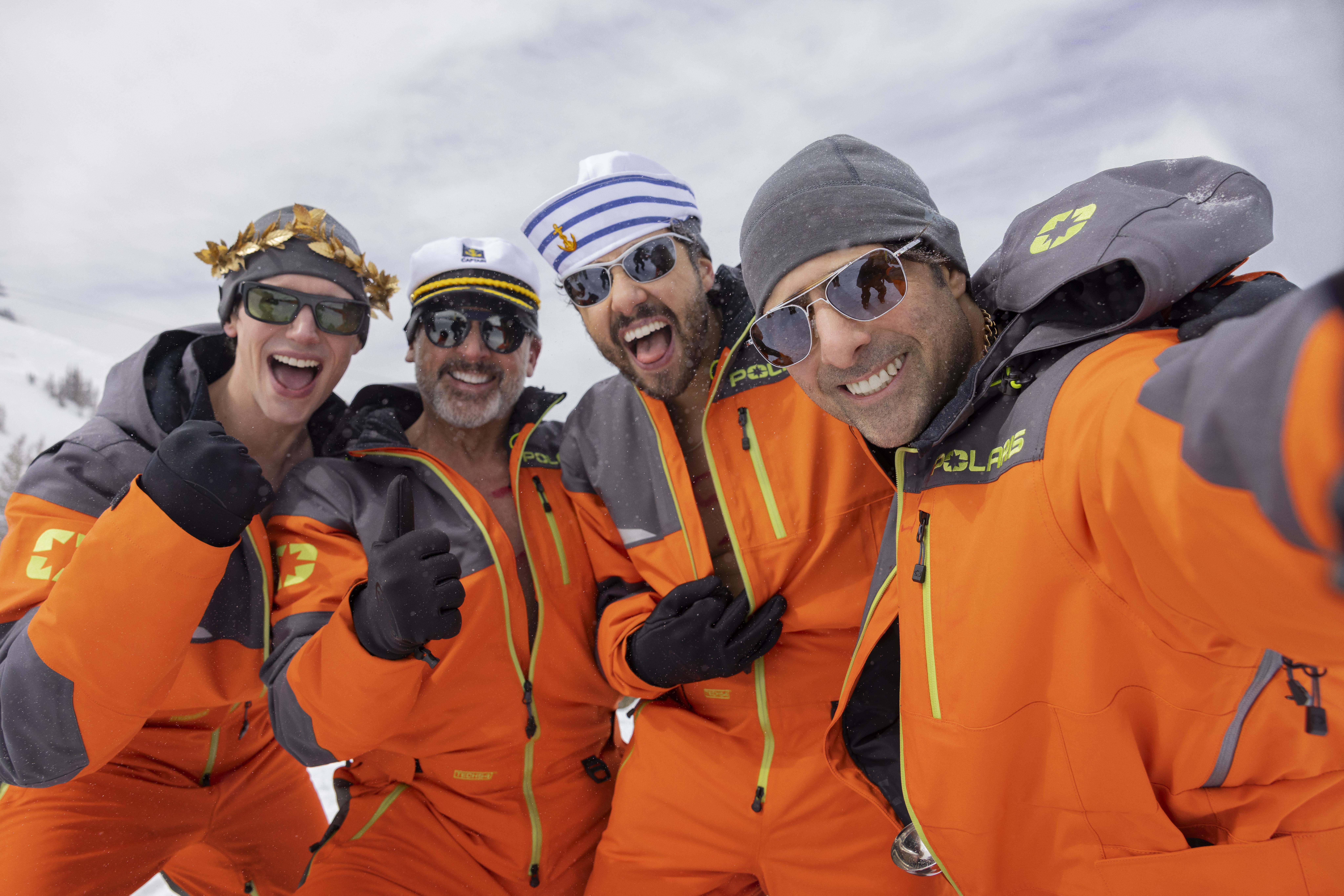 Four men in orange snowsuits and playful headwear give thumbs up.