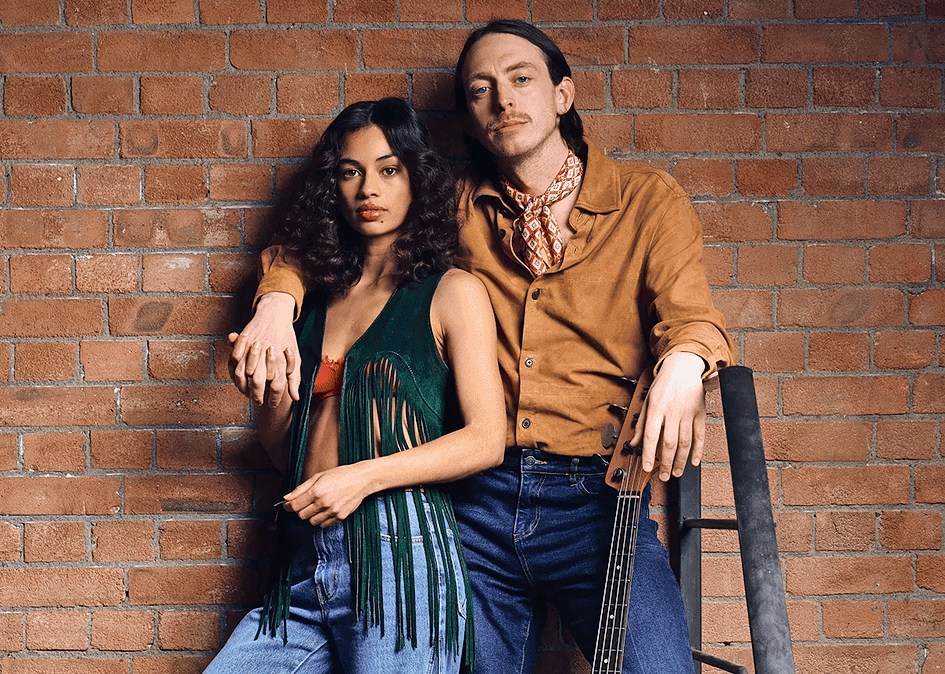 Nia Towle and Zachary Hart, in 70s attire, leaning against a brick wall with a bass guitar.